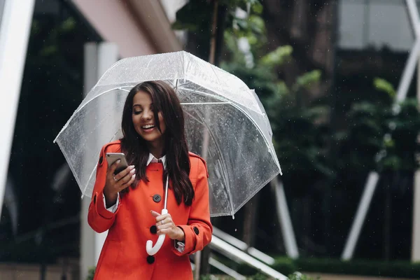 Portret van de vrouw in het rood met transparante paraplu lezen goed nieuws in een slimme telefoon in de stad. — Stockfoto