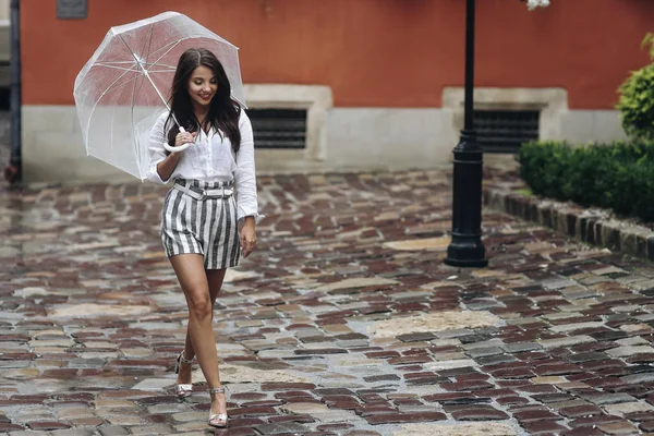 Modelo bonito atravessa a cidade a pé. Linda morena jovem vestindo shorts, andando na rua com guarda-chuva transparente . — Fotografia de Stock