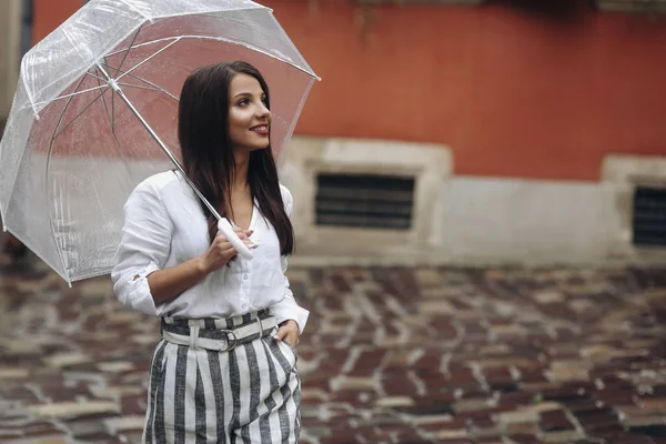 Schöne junge Frau, die durch die Stadt geht. schöne brünette junge Frau trägt weißes Hemd, geht auf der Straße mit durchsichtigem Regenschirm. — Stockfoto