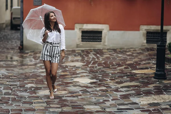 街を歩き回るカジュアルな服を着た美しいブルネットの女の子、ファッションスタイル。美しいブルネットの若い女性は、透明な傘で通りを歩いて、ショートパンツを着て. — ストック写真