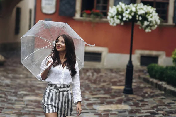 Junges schönes Mädchen, das durch die Stadt geht. schöne brünette junge Frau in kurzen Hosen, die mit durchsichtigem Regenschirm auf der Straße spaziert. — Stockfoto