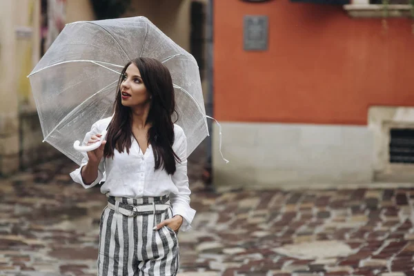 Een wandeling in het centrum van de stad, vrouw in wit shirt wandelingen rond de stad. Mooie brunette jonge vrouw het dragen van wit shirt, wandelen op de straat met transparante paraplu. — Stockfoto