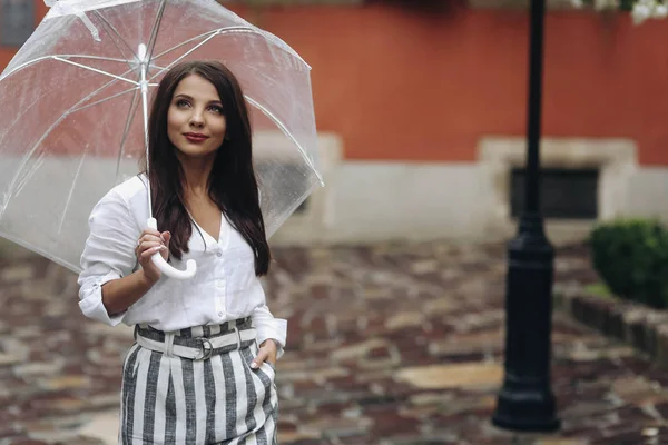 Un paseo por el centro de la ciudad, una mujer joven caminando por la ciudad. Hermosa morena joven con camisa blanca, caminando por la calle con paraguas transparente . — Foto de Stock