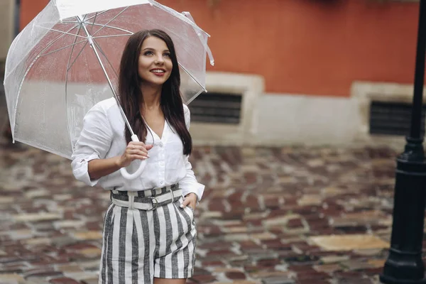 Una mujer joven y sonriente posando en la cámara de la ciudad. Hermosa morena joven con camisa blanca, caminando por la calle con paraguas transparente . — Foto de Stock