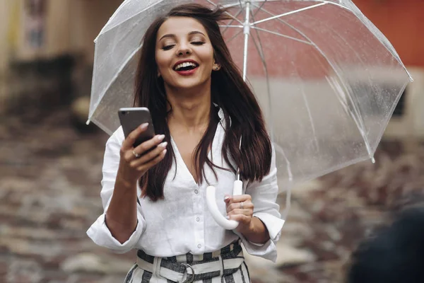 Portret van lachen mooi meisje met paraplu noemt een vriend die op de oude straat staat. Het is zomer regen. — Stockfoto