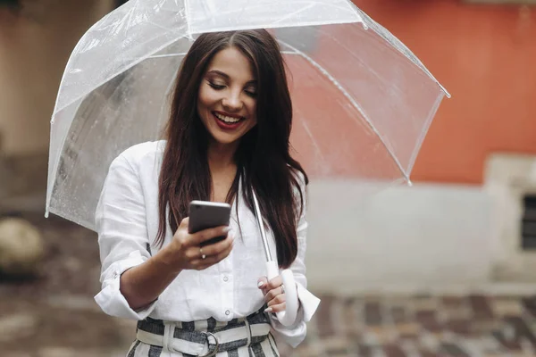 Portret van glimlachend mooi meisje met paraplu. met behulp van smartphone, rode huis op de achtergrond. Het is zomer regen. — Stockfoto