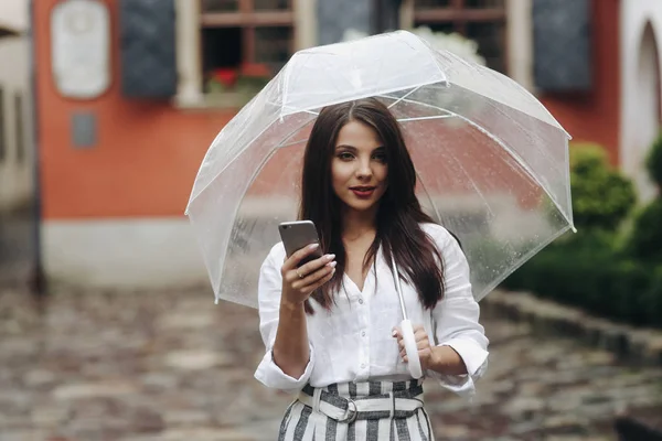 Portret van dromerig mooi meisje met paraplu, met behulp van smartphone, rode huis op de achtergrond. Kleine scherptediepte. — Stockfoto