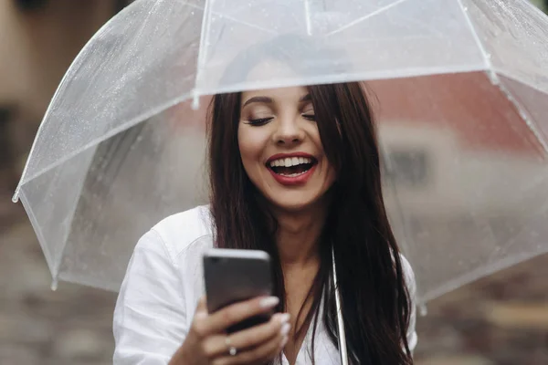 Закройте портрет улыбающейся красивой девушки с пуговицей, используя смартфон, красной стеной на заднем плане. Небольшая глубина резкости. Летний дождь. . — стоковое фото