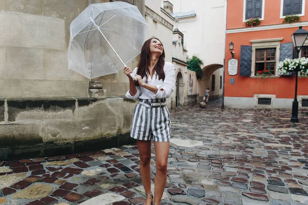 Un paseo por el centro de la ciudad, mujer joven y de moda caminando por la ciudad, expresión facial. Hermosa morena joven con pantalones cortos, caminando por la calle con paraguas transparente . — Foto de Stock