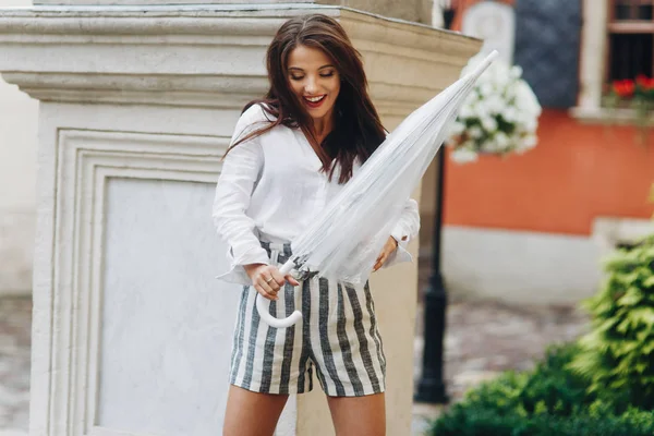 Retrato de una chica emocional jugando con su paraguas, de pie cerca de la columna. Hermosa morena joven con pantalones cortos, caminando por la calle con paraguas transparente . — Foto de Stock