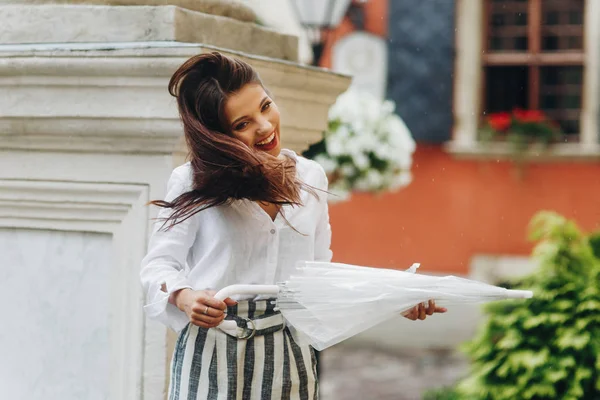 Chica morena con estilo caminando por la ciudad, jugando con su paraguas, de pie cerca de la columna. Hermosa joven morena caminando por la calle con paraguas transparente . — Foto de Stock