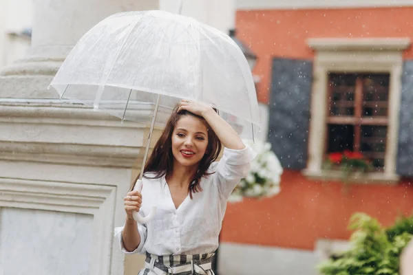 Elegante vrouw wandelingen rond de stad onder paraplu. Brunette jonge vrouw het dragen van wit shirt, wandelen op de straat met transparante paraplu. — Stockfoto