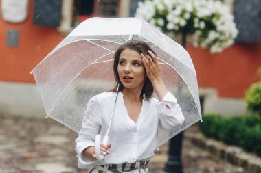 Genç bir kadın şehirde şemsiyeyle dolaşıyor, yaz yağmuru yağıyor. Beyaz gömlek giyen esmer genç kadın, şeffaf şemsiye ile sokakta yürüyor.