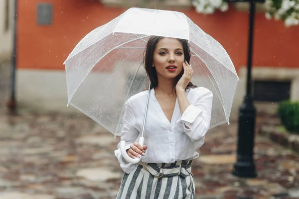 Beautiful girl dreamily walking around cozy city street with umbrella, casual style. It\'s summer rain.