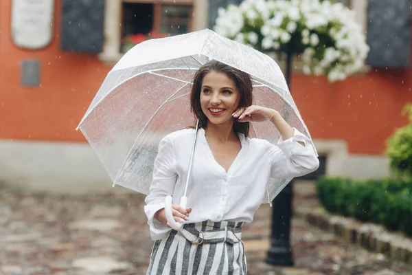 Eine junge Frau läuft mit Regenschirm und lässigem Stil durch die Stadt. Es ist Sommerregen. brünette junge Frau in weißem Hemd, mit durchsichtigem Regenschirm auf der Straße. — Stockfoto