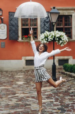 Muhteşem genç kadın şehirde dolaşırken gülümsüyor, şemsiyesiyle oynuyor. Yaz yağmuru..