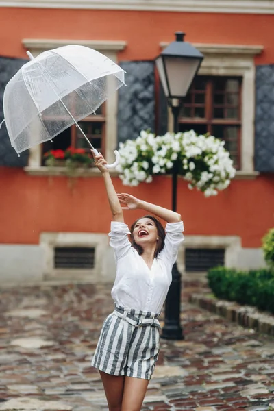 Elegante vrouw loopt rond de stad, spelen met haar paraplu. Het is zomer regen. Brunette jonge vrouw dragen shirt en shorts, wandelen op de straat met transparante paraplu. — Stockfoto