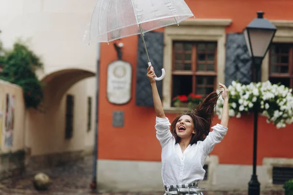 Mujer positiva y hermosa paseando por la ciudad verano. Es lluvia de verano. Mujer joven morena con camisa blanca, caminando por la calle con paraguas transparente . — Foto de Stock