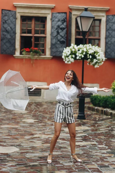 Begreppet liv modern kvinna i storstad, leker med sitt paraply. Det är sommarregn. Brunett ung kvinna klädd i skjorta och shorts, gå på gatan med transparent paraply. — Stockfoto