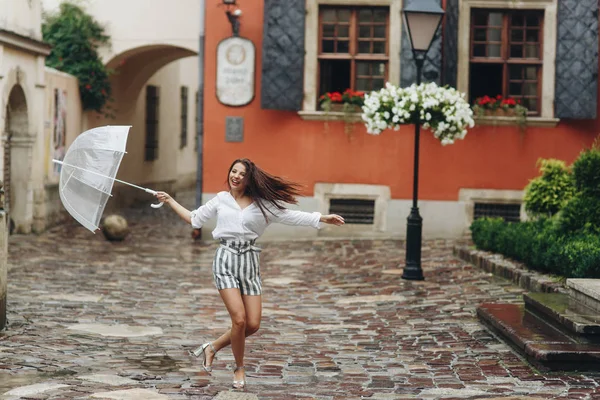 Hermosa chica vestida a la moda caminando por la ciudad cerca del antiguo edificio, jugando con su paraguas. Es lluvia de verano. . — Foto de Stock