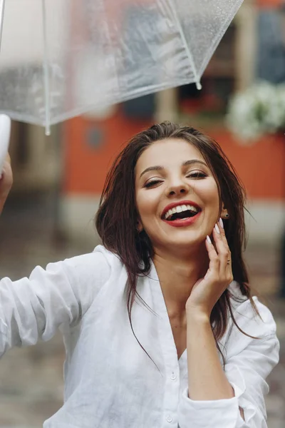 Vertikal porträtt av en positiv och vacker kvinna njuter sommardag under helgen. Brunett ung kvinna klädd i vit skjorta, gå på gatan med transparent paraply. — Stockfoto