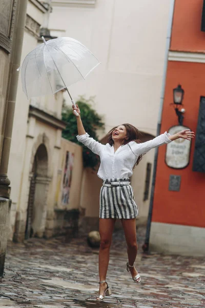 Vertikal porträtt av en lycklig flicka med paraply nära gamla hus hoppar i luften. Kopiera utrymme. Det är sommarregn. — Stockfoto