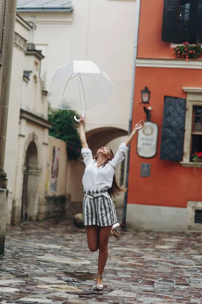 Porträtt av en lycklig flicka med paraply nära gammalt hus. Det är sommarregn. Brunett ung kvinna klädd i vit skjorta, gå på gatan med transparent paraply. — Stockfoto
