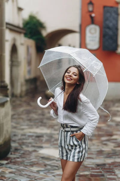 Portret van een lachende gelukkige vrouw onder een paraplu in de buurt van het oude huis. Kopieer ruimte. Het is zomer regen. Brunette jonge vrouw dragen shirt en shorts, hand in de zak — Stockfoto
