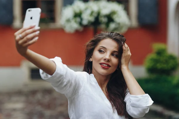 Горизонтальный портрет довольно счастливой улыбающейся молодой женщины в белой рубашке, делающей селфи с мобильным телефоном на фоне дома с цветами . — стоковое фото