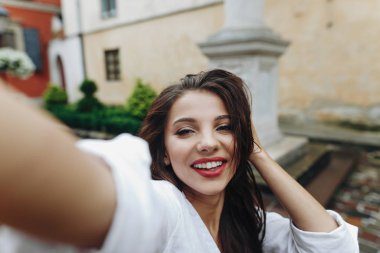 Yakın yatay oldukça mutlu güzel gülümseyen genç kadın sokak arka planda cep telefonu ile selphie yapma ve saçlarını sabitleme.