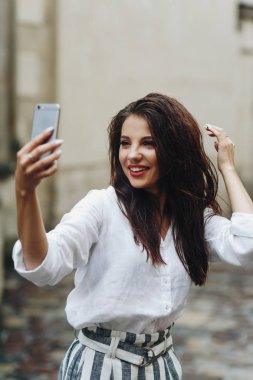 Güzel esmer kız yaz günü poz. Eski beyaz duvarın arka planında cep telefonu ile selphie yapan gülümseyen genç kadının dikey portresi.