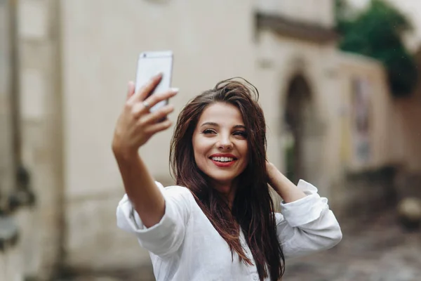 Beautiful lovely smiling young brunette woman wearing white shirt making self portrait with mobile phone on the background of old house. — Stock Photo, Image