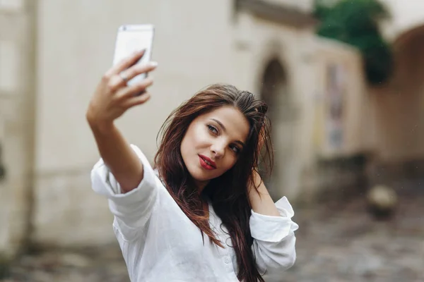 Путешествует красивая молодая брюнетка женщина в белой рубашке делает автопортрет с мобильного телефона на фоне старого дома — стоковое фото