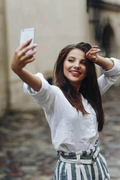 Позирование в летний день. Вертикальный портрет улыбающейся молодой женщины, делающей селфи с помощью мобильного телефона на фоне старого белого дома . — стоковое фото