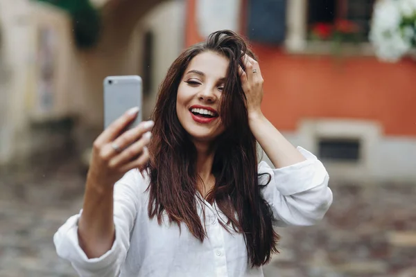 Горизонтальный портрет довольно счастливой милой улыбающейся молодой женщины в белой рубашке, использующей телефон для интернет-звонка в городе . — стоковое фото