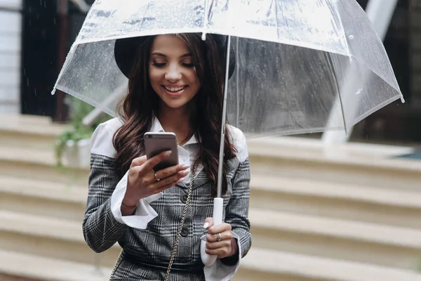 Портрет красивой смеющейся женщины в черной шляпе и сером пальто под прозрачным зонтиком, стоящей на улице, беседующей с друзьями. Хорошие новости . — стоковое фото