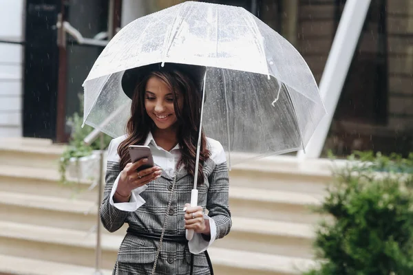Retrato de una hermosa mujer sonriente vestida de negro ha? t y abrigo gris bajo paraguas transparente de pie en la calle, escribiendo sms . — Foto de Stock