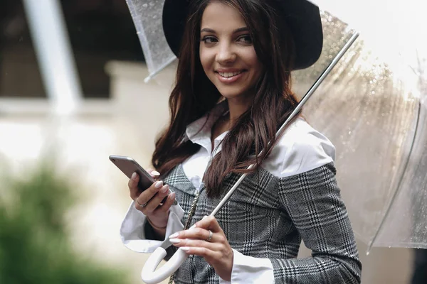 Primer plano retrato de hermosa mujer sonriente con sombrero negro y abrigo gris bajo paraguas transparente, escribiendo sms — Foto de Stock