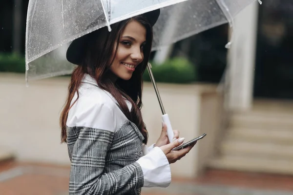 Retrato de una hermosa mujer con sombrero negro y abrigo gris bajo paraguas transparente, sosteniendo el teléfono inteligente en su mano —  Fotos de Stock
