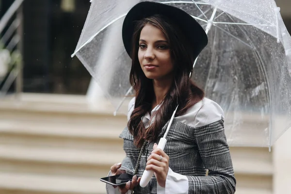 Nahaufnahme Porträt einer schönen Frau mit schwarzem Hut und grauem Mantel unter transparentem Regenschirm, Smartphone in der Hand — Stockfoto