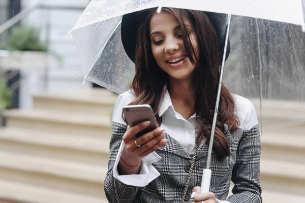 Close up portrait of beautiful smiling woman wearing black hat and gray coat under transparent umbrella — Stock Photo, Image