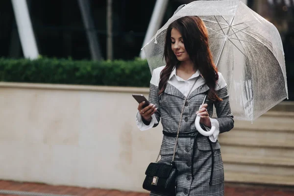 Portret van mooie vrouw in afwachting van het ontmoeten van haar vriendje. Vrouw dragen grijze vacht onder paraplu, het houden van smartphone in uw hand. Het is lente regen. — Stockfoto