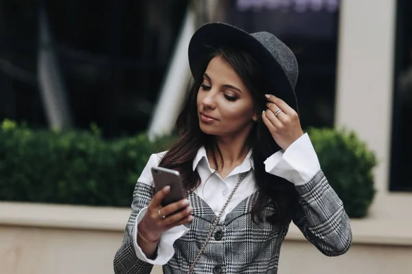 Портрет красивой серьезной женщины в черной шляпе и серой куртке, стоящей возле кафе, читающей смс . — стоковое фото