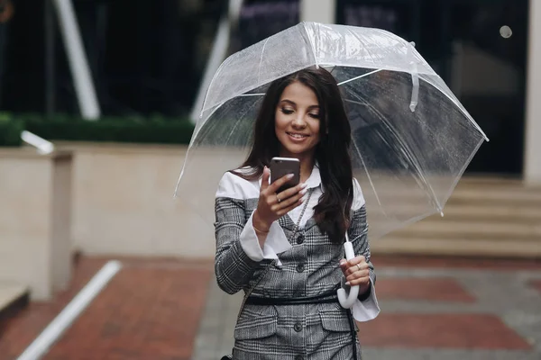 Портрет красивой улыбающейся женщины со смартфоном в руке и печатанием смс. Женщина в сером пальто под зонтиком получила смешное SMS-сообщение — стоковое фото