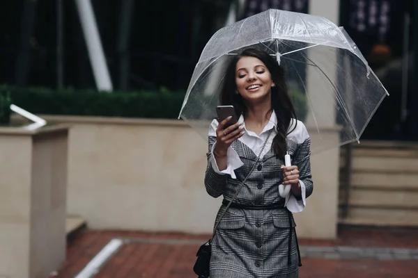 Porträt einer schönen lächelnden Frau mit Smartphone in der Hand und tippenden SMS in der Nähe eines Cafés. Frau mit grauem Mantel unter Regenschirm — Stockfoto