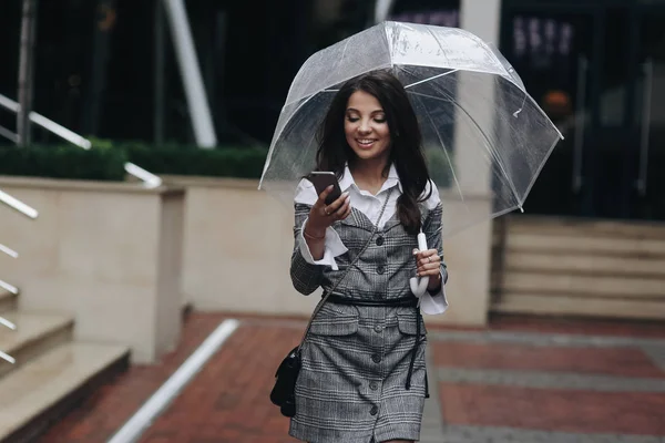 Porträtt av vacker kvinna med smartphone i hand och skriva SMS stående nära kontoret. Kvinna klädd i grått skikt under paraply fick en rolig SMS — Stockfoto
