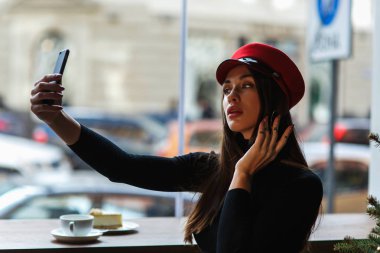 Sakin genç kadın akıllı telefonuyla selfie çekiyor. Şehrin arka planda kafede oturan kırmızı şapkalı moda modeli nin yakın portresi. O rahat kafede otururken poz