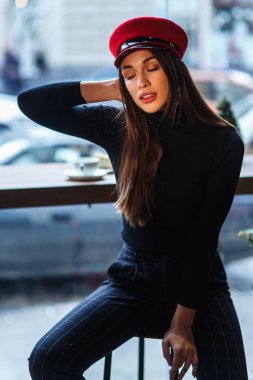 Gözleri kapalı kafede sandalyede oturan kırmızı şapkalı romantik kadın portresi. Fotokopi alanı ile şehrin arka planına karşı kafede Kadın. Güzellik ve moda kavramı
