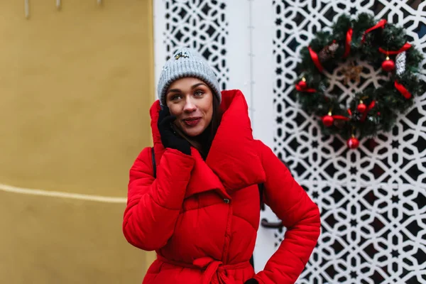 年轻女子在墙的背景与圣诞花圈在手机上交谈 冬天那天 穿着红色冬衣的女人站在大街上 圣诞假期概念与复制空间 — 图库照片