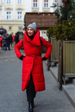 Noel ağaçları satış yeri yakınında sokakta güzel kadının tam uzunlukta sokak portresi. Soğuk kış gününde sokakta yürüyen kırmızı kış ceketli kadın. Noel tatili konsepti